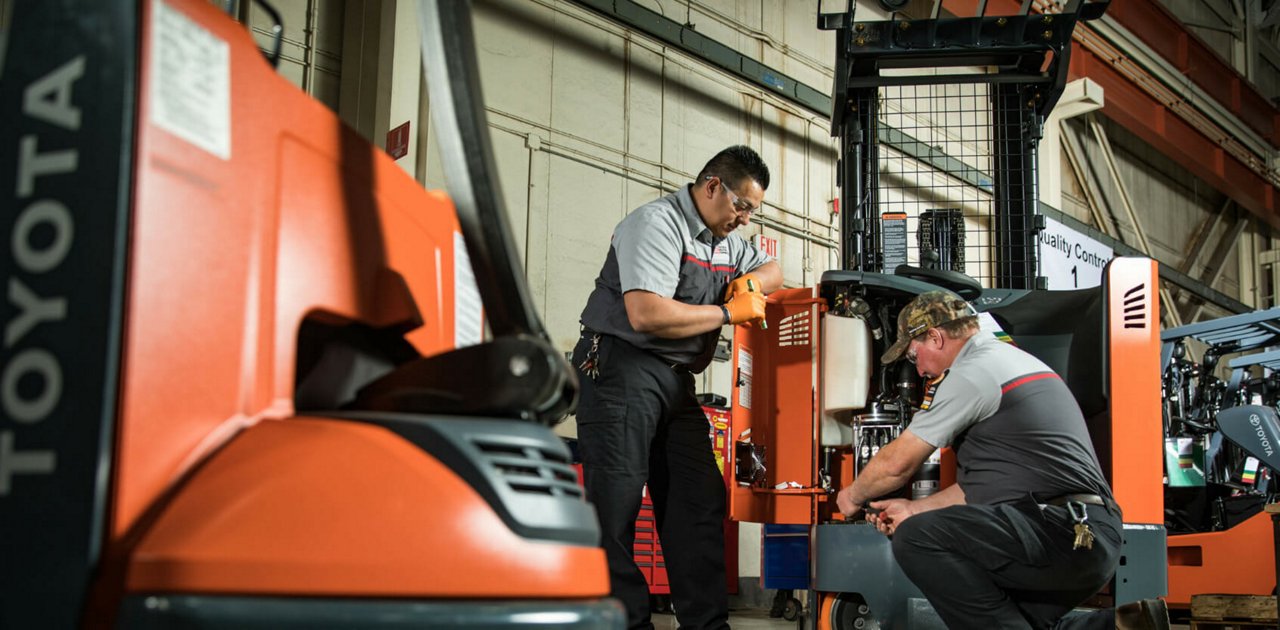 Two men talking in a warehouse while one works on a forklift and the other is standing by 