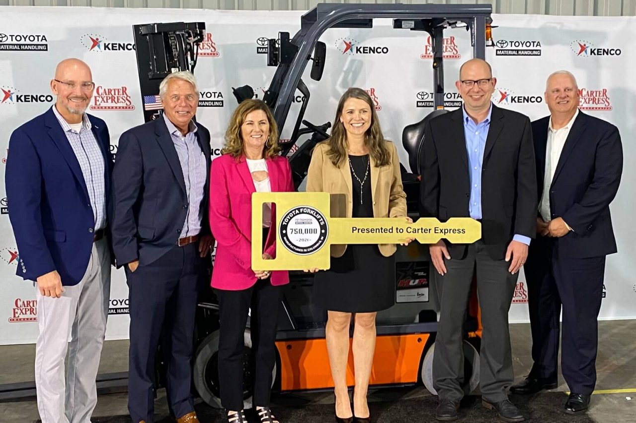 4 men and 2 women carrying a large key as an award, infront of a photo station and a forklift 