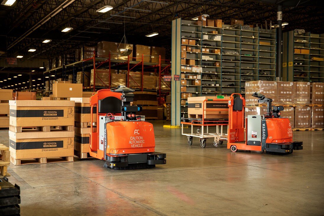 Two automated forklifts in a warehouse, one driving with a cardboard box on its pallet and one parked