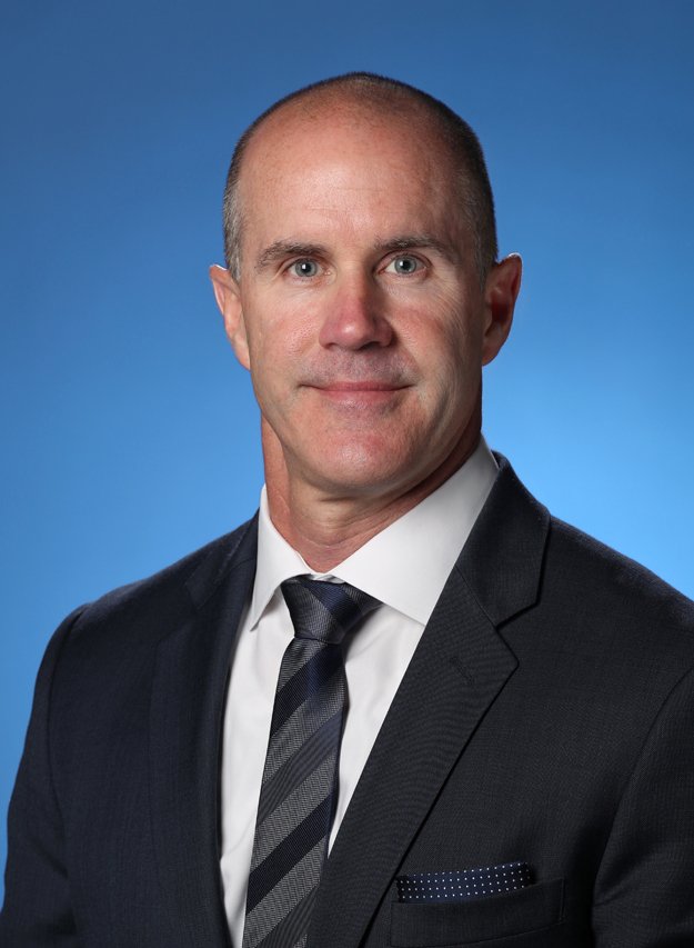 headshot of middle aged white male in a black suit jacket and black tie against a blue background