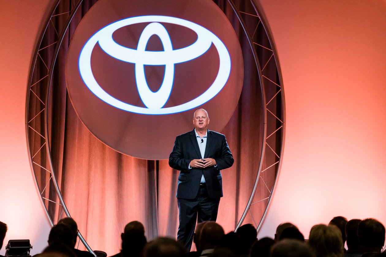 Bret Bruin speaking at a conference on stage to a crowd of people, with the Toyota logo as a backdrop behind him