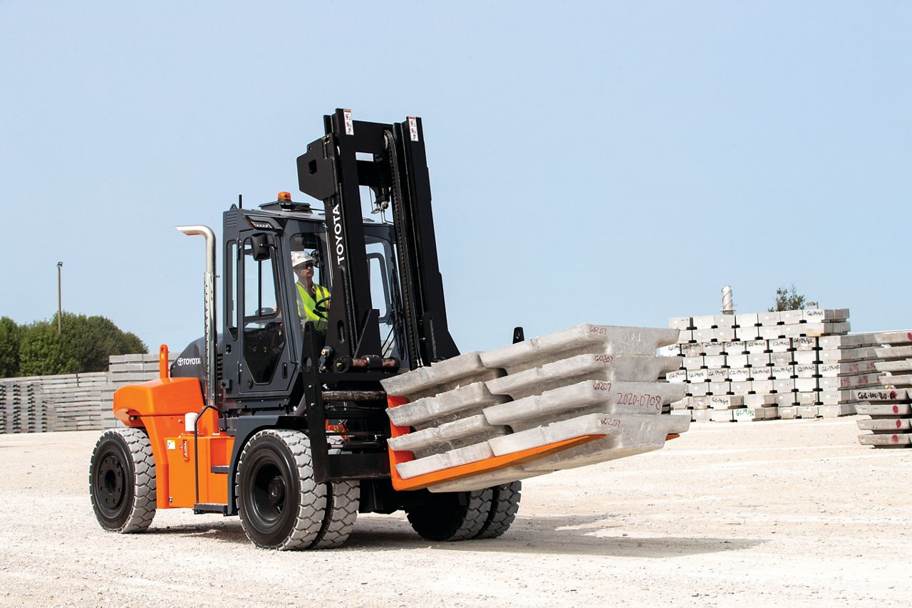 A man driving a forklift carrying a pallet of 8 building blocks in a freight yard outside. 