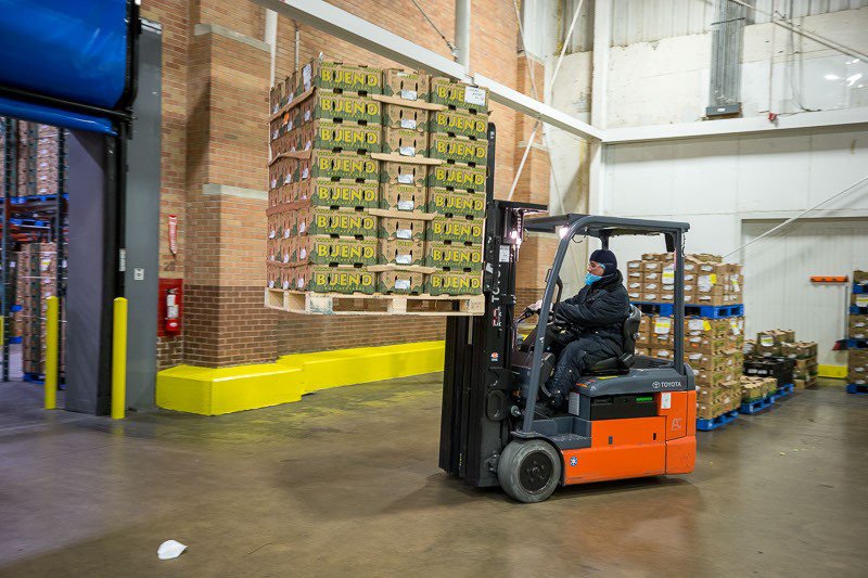 A man driving a forklift in a food storage warehouse, lifting a pallet with many cardboard boxes of food. 