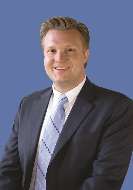 Portrait professionnel d’un homme blanc aux cheveux blonds portant une veste de costume noire et une cravate bleue sur fond bleu