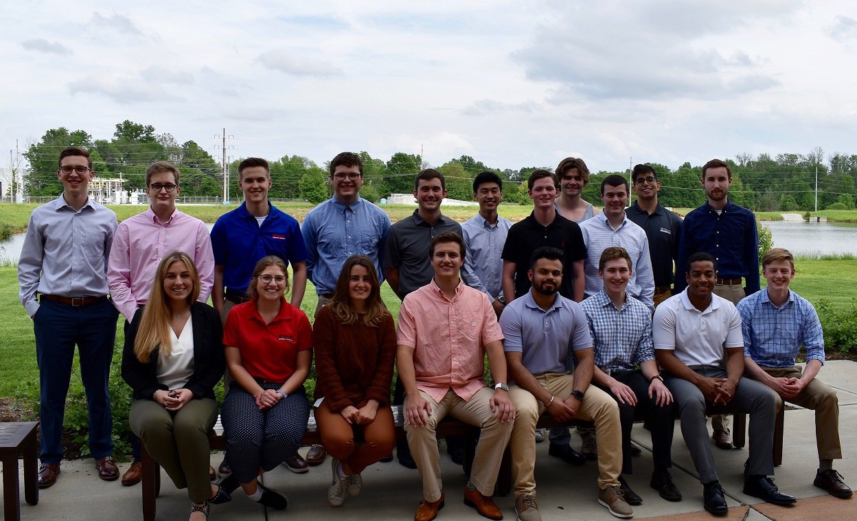 8 interns are sitting on a bench outside, with 11 interns standing behind them