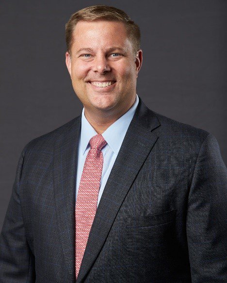 headshot of man in black suit jacket and red tie against a grey background