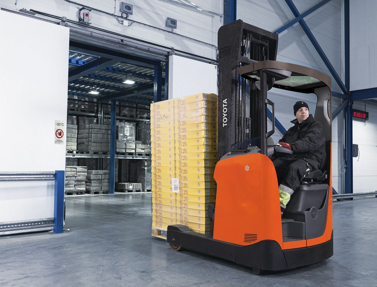 Homme portant un manteau épais et un chapeau tout en utilisant un chariot élévateur orange dans un environnement d’entreposage frigorifique