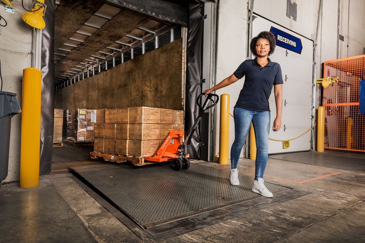 Woman Using A Hand Pallet Truck Transporting Cardboard Boxes In The Warehouse 