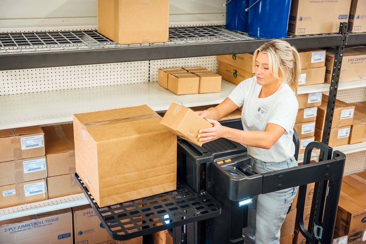 Trabajadora de almacén rubia con camiseta blanca y jeans cargando cajas en la parte superior de la bandeja de carga del recogedor de existencias