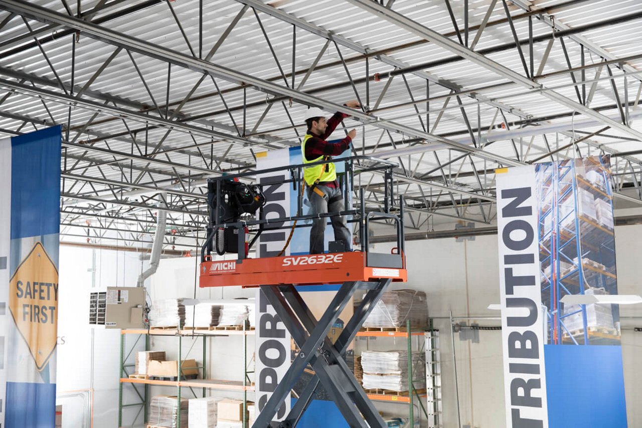 Warehouse worker using an Aichi Scissor Lift to adjust a fixture on the ceiling of a warehouse