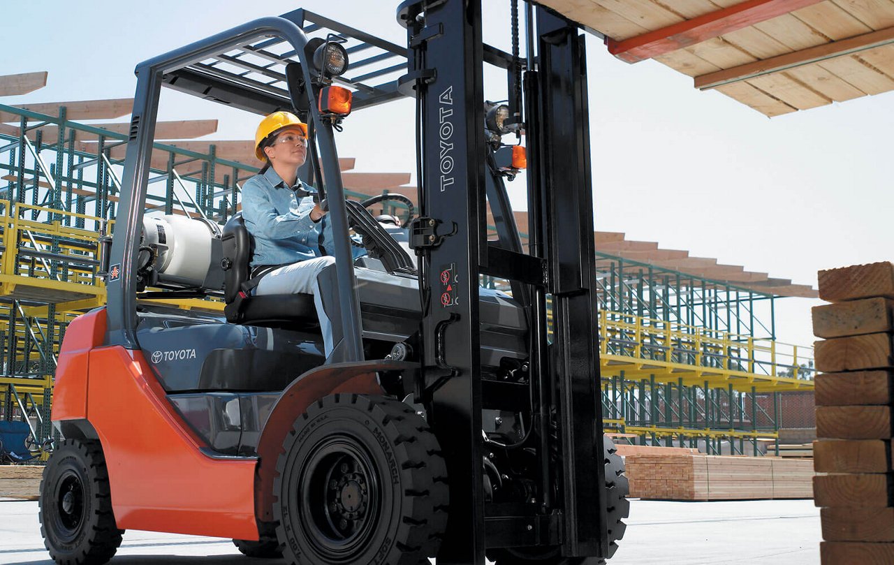 A woman driving a Core IC Pneumatic forklift in an outdoor loading dock to carry a wooden pallet 