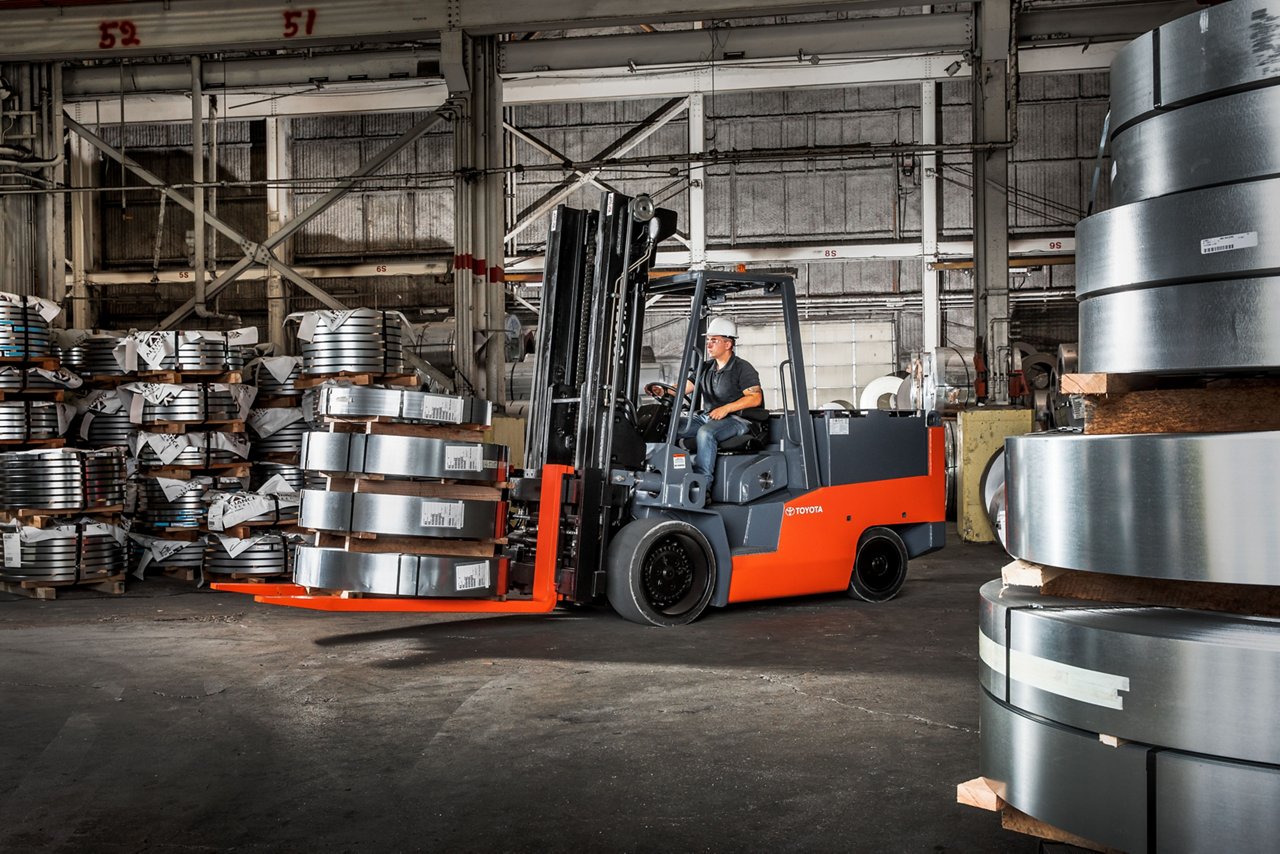 Man driving Toyota High capacity electric cushion forklift and carrying 6 large metal rings in warehouse. 