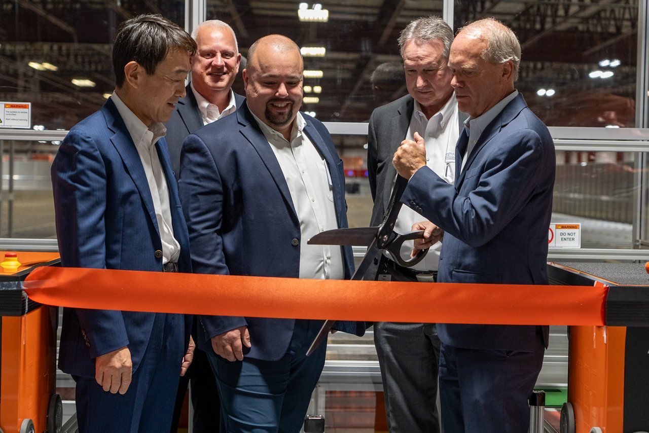 Toyota and Bastian Solutions executives stand in front of orange ribbon as Brett Wood, President & CEO of TMHNA holds al large pair of scissors to cut the ribbon