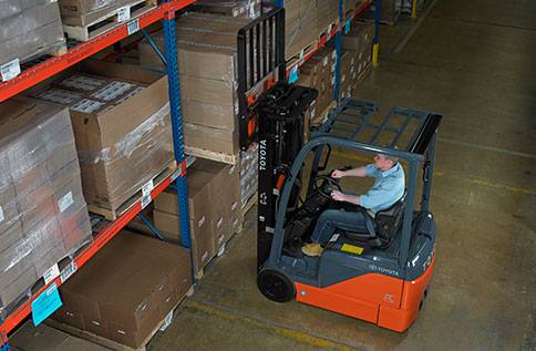 Man driving a Toyota forklift in a warehouse, lifting large cardboard box to high shelf 