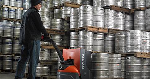 Man using Toyota Pallet Jack to carry 2 metal barrels in warehouse